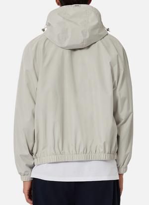 A person is seen from behind wearing Industrie The Rain Jacket in light gray, featuring a drawstring hood and elastic waistband. A white shirt underneath subtly contrasts with dark pants. The jacket is branded with the Industrie logo.