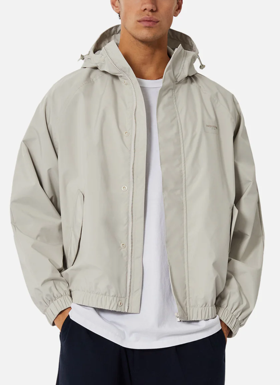 Someone is dressed in a casual Industrie The Rain Jacket, light gray with a hood, worn over a white t-shirt and black pants. The jacket, partially zipped with hands in pockets, sports an Industrie logo on the front against a plain white backdrop.