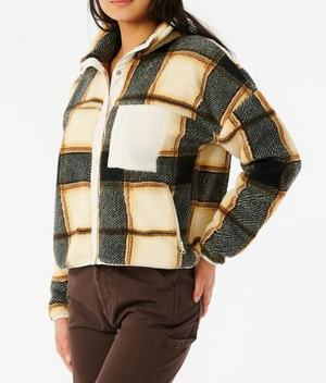 A person wears the Rip Curl Kindred Palms Polar Fleece—an oversized fuzzy plaid jacket with beige, black, and brown checks plus a white chest pocket—paired with brown pants, standing against a white background.