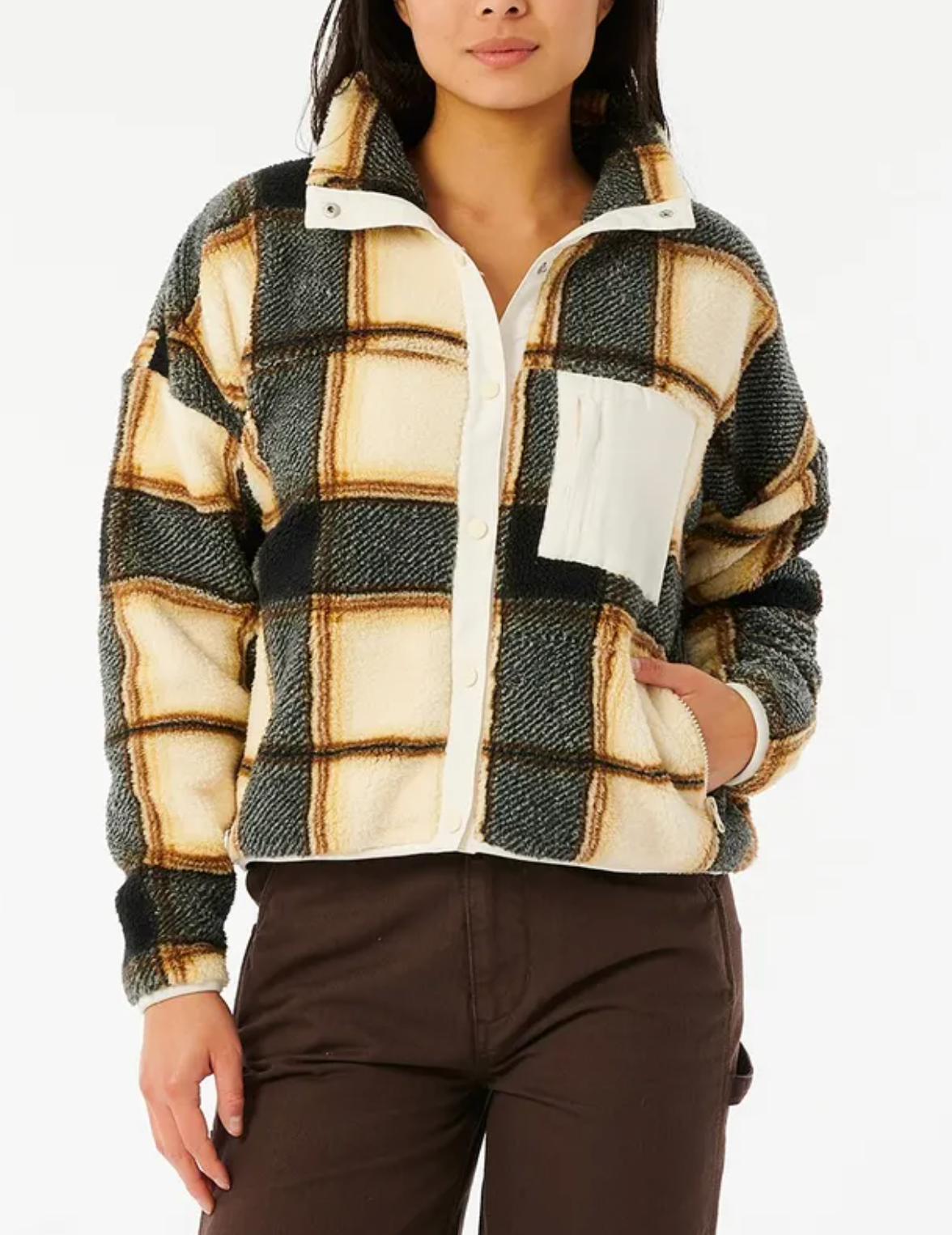 A person wears the Rip Curl Kindred Palms Polar Fleece—an oversized cream, black, and tan plaid jacket with a white chest pocket—paired with brown pants, standing against a plain white background.