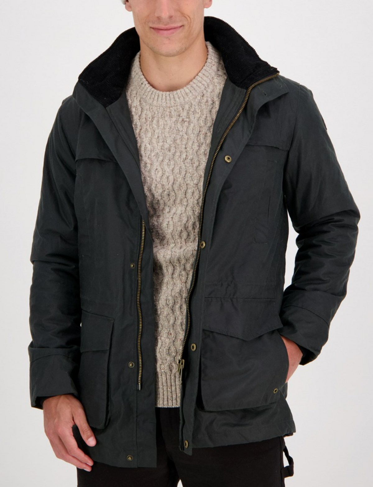 Wearing a Swanndri Evans Flat Oilskin Jacket over a light knitted sweater, a person stands against a white background. The open jacket features large pockets and a textured collar, with hands tucked inside, exuding rustic charm and practicality reminiscent of classic Swanndri style.
