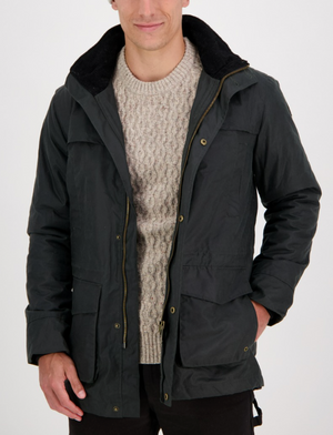 Wearing a Swanndri Evans Flat Oilskin Jacket over a light knitted sweater, a person stands against a white background. The open jacket features large pockets and a textured collar, with hands tucked inside, exuding rustic charm and practicality reminiscent of classic Swanndri style.
