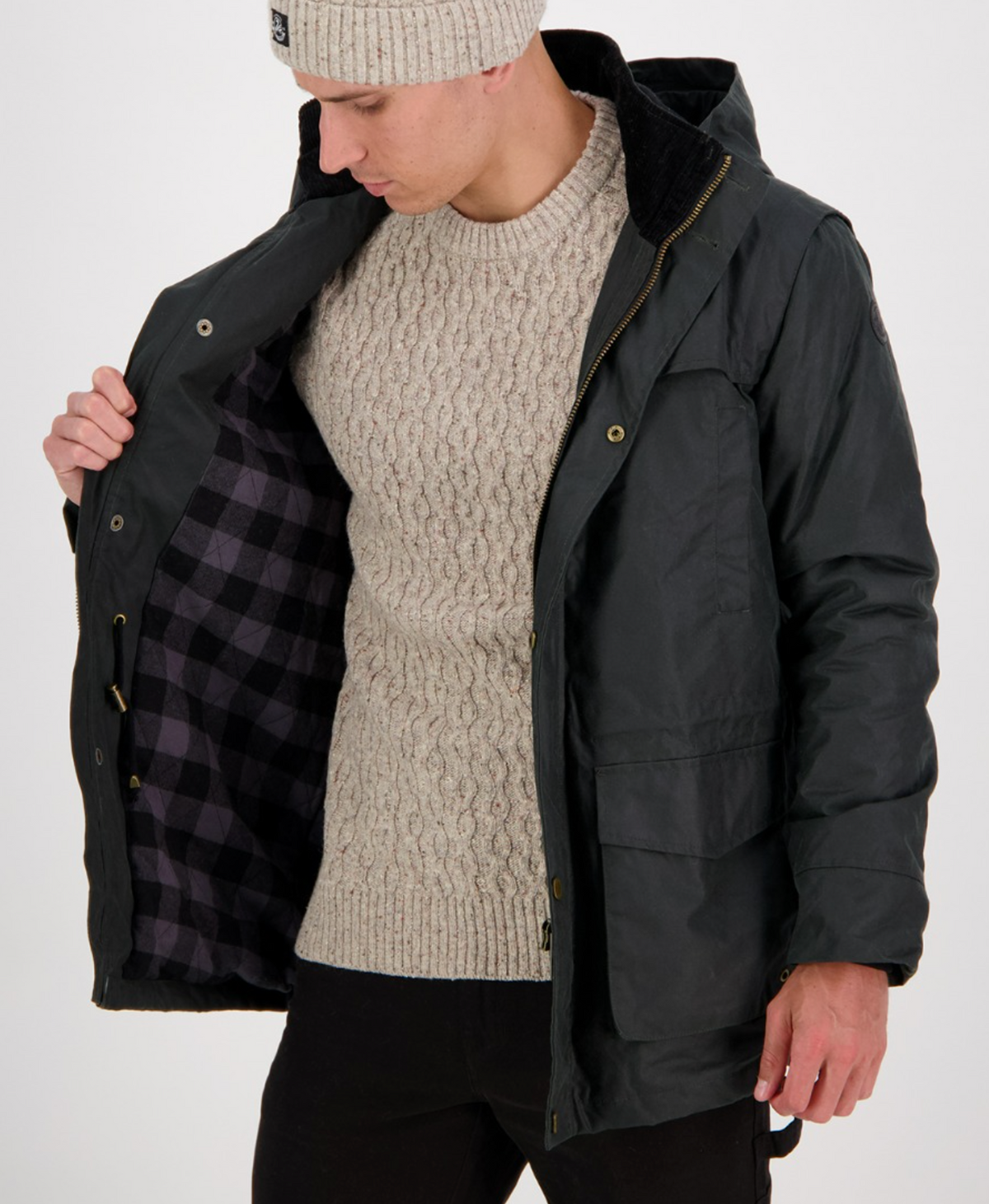 Wearing black pants and the Swanndri Evans Flat Oilskin Jacket in dark green, unzipped to show a plaid lining, a person tops off their outfit with a beige knit sweater and matching beanie as they gaze downward against a plain white background.