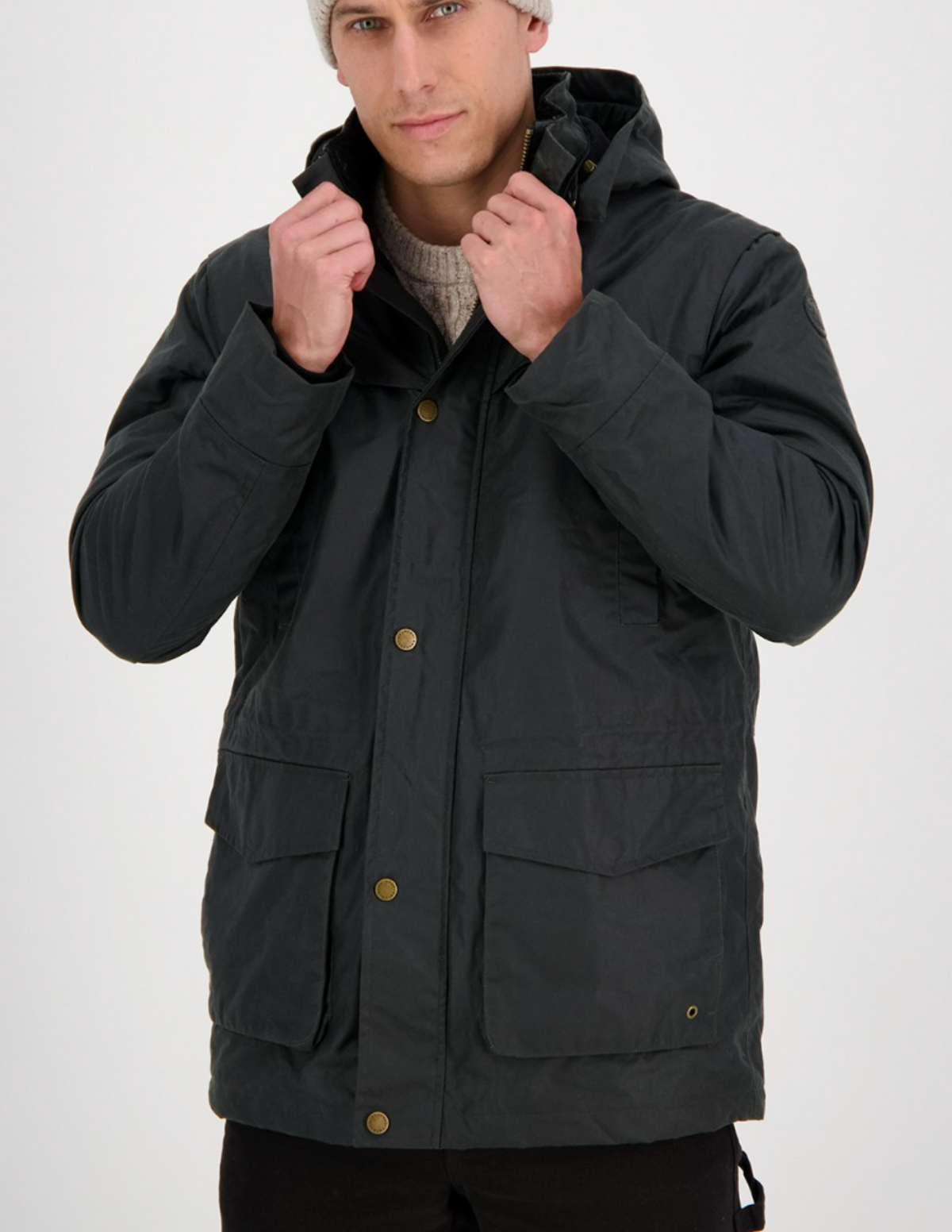 A man in a dark Swanndri Evans Flat Oilskin Jacket with large front pockets and brass buttons adjusts the collar. The detachable hood is tucked away, complementing his light-colored knitted beanie and sweater underneath, all set against a plain white background that highlights his rugged style.