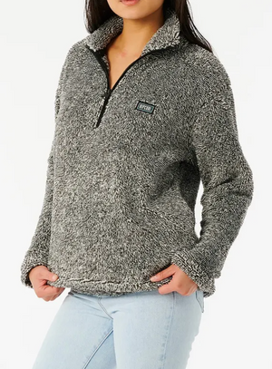 A person in a Rip Curl Dark N Stormy II cozy, textured gray half-zip pullover with a small logo on the chest has their hands in the pockets while wearing light blue jeans. The image is cropped at the neck and waist.
