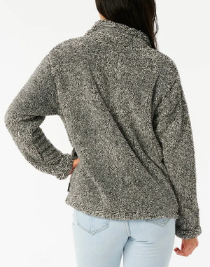 A person with long hair is wearing a cozy Rip Curl Dark N Stormy II half-zip gray sweater and light blue jeans. The person faces away from the camera, one hand on their hip, against a plain white background.