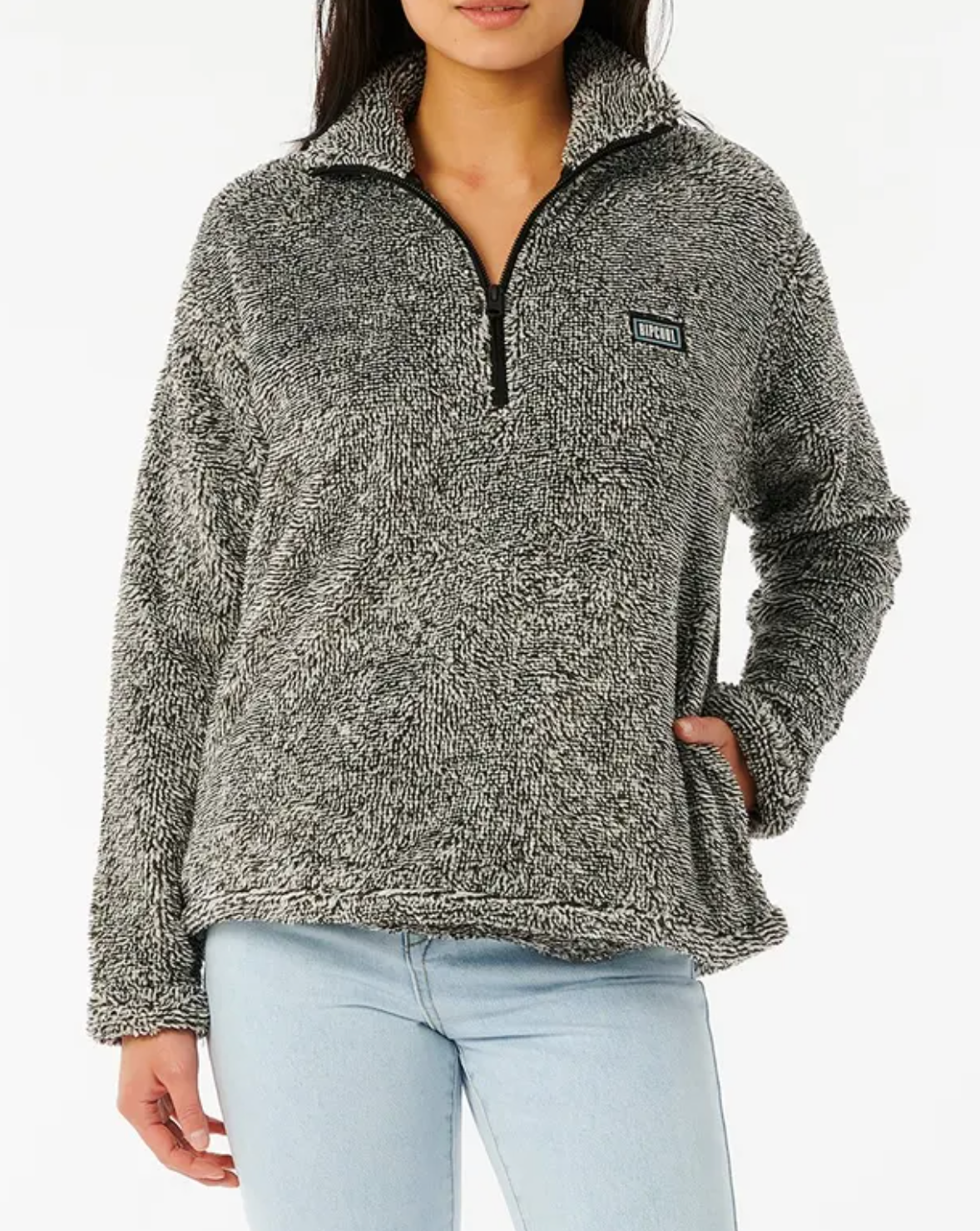 A person in a cozy Rip Curl Dark N Stormy II gray fleece pullover with a half zip and chest patch, paired with light blue jeans and one hand in their pocket against a plain white background.