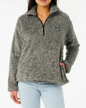 A person in a cozy Rip Curl Dark N Stormy II gray fleece pullover with a half zip and chest patch, paired with light blue jeans and one hand in their pocket against a plain white background.