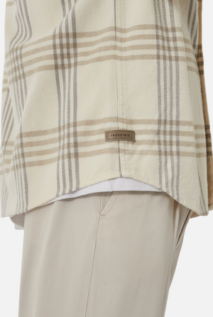 Close-up of a person wearing the Industrie The Danville Shirt, a regular fit cream plaid shirt made from 100% cotton, with tan and gray stripes over beige pants and a small "INDUSTRIE" tag near the hem.