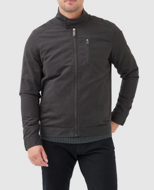 A person wearing the stylish Rodd & Gunn Armitage Jacket, which features a front zipper and a small zippered pocket on the chest, has their hands in the jacket pockets. The individual, with short hair, is dressed in black pants and a gray shirt underneath this comfortable jacket made of Australian cotton. The background is plain white.
