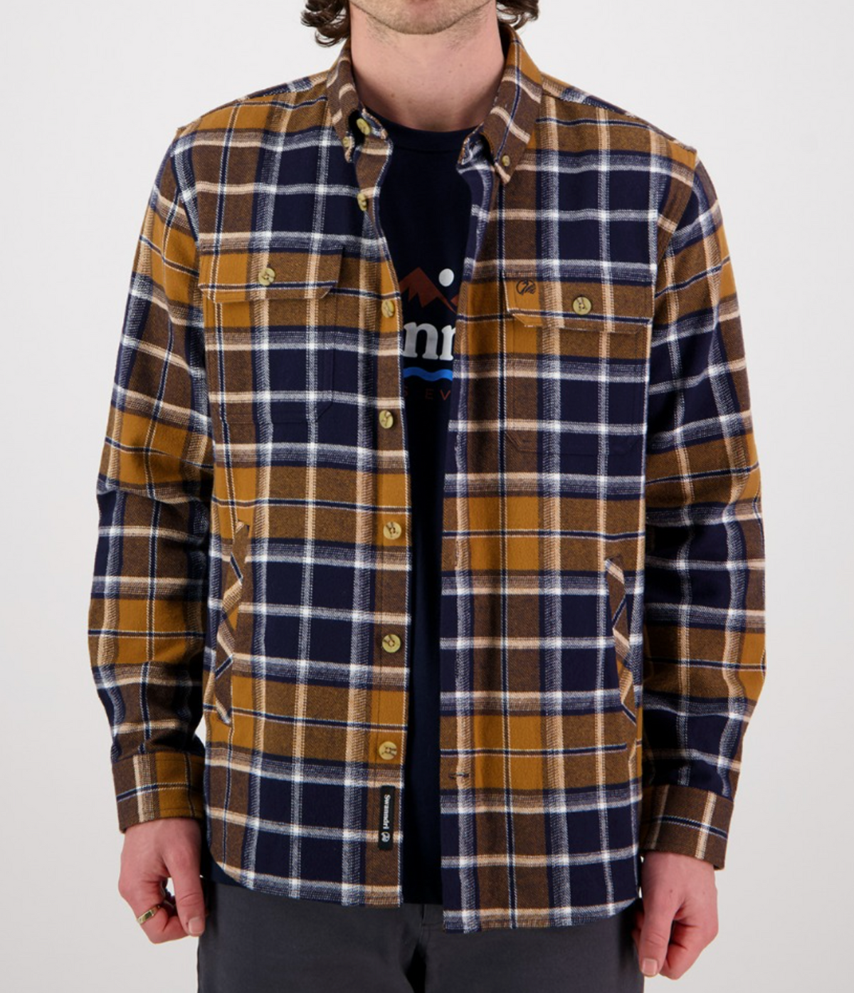 A person wearing the Swanndri Pencarrow Shacket—a 100% cotton, classic fit plaid shacket in mustard yellow, navy blue, and white—over a dark graphic tee with gray pants stands facing the camera against a plain background.