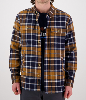 A person wearing the Swanndri Pencarrow Shacket—a 100% cotton, classic fit plaid shacket in mustard yellow, navy blue, and white—over a dark graphic tee with gray pants stands facing the camera against a plain background.