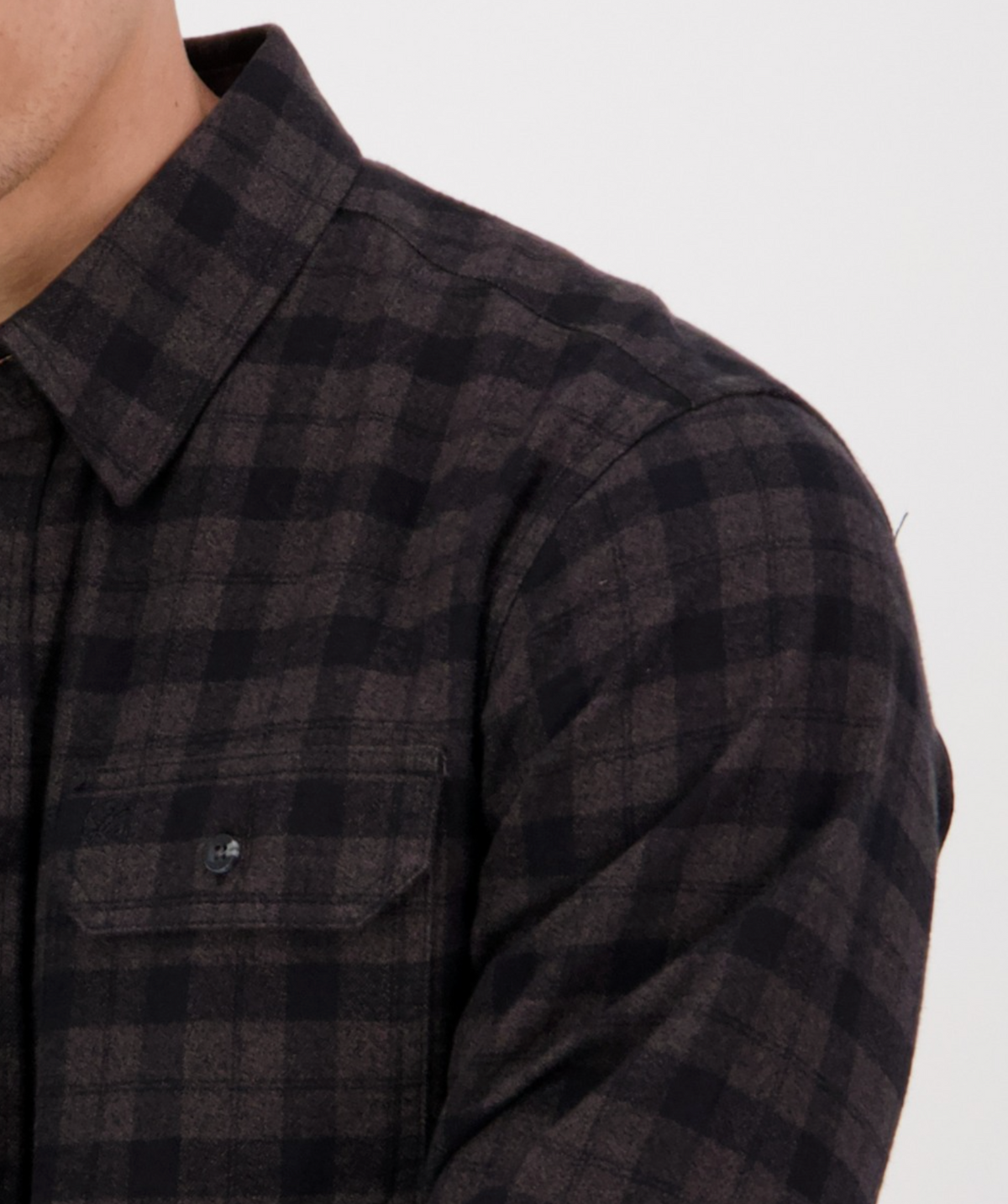 Close-up of someone in a Swanndri Port Road Shirt, 100% cotton with black and dark gray plaid twill. It has a buttoned collar and a visible chest pocket. Only part of the person's face is seen against a plain white background.