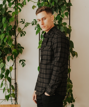 In front of a wall of green plants, a person wears the Swanndri Port Road Shirt—a black and gray checkered style with a subtle twill design—paired with black pants, looking towards the camera.