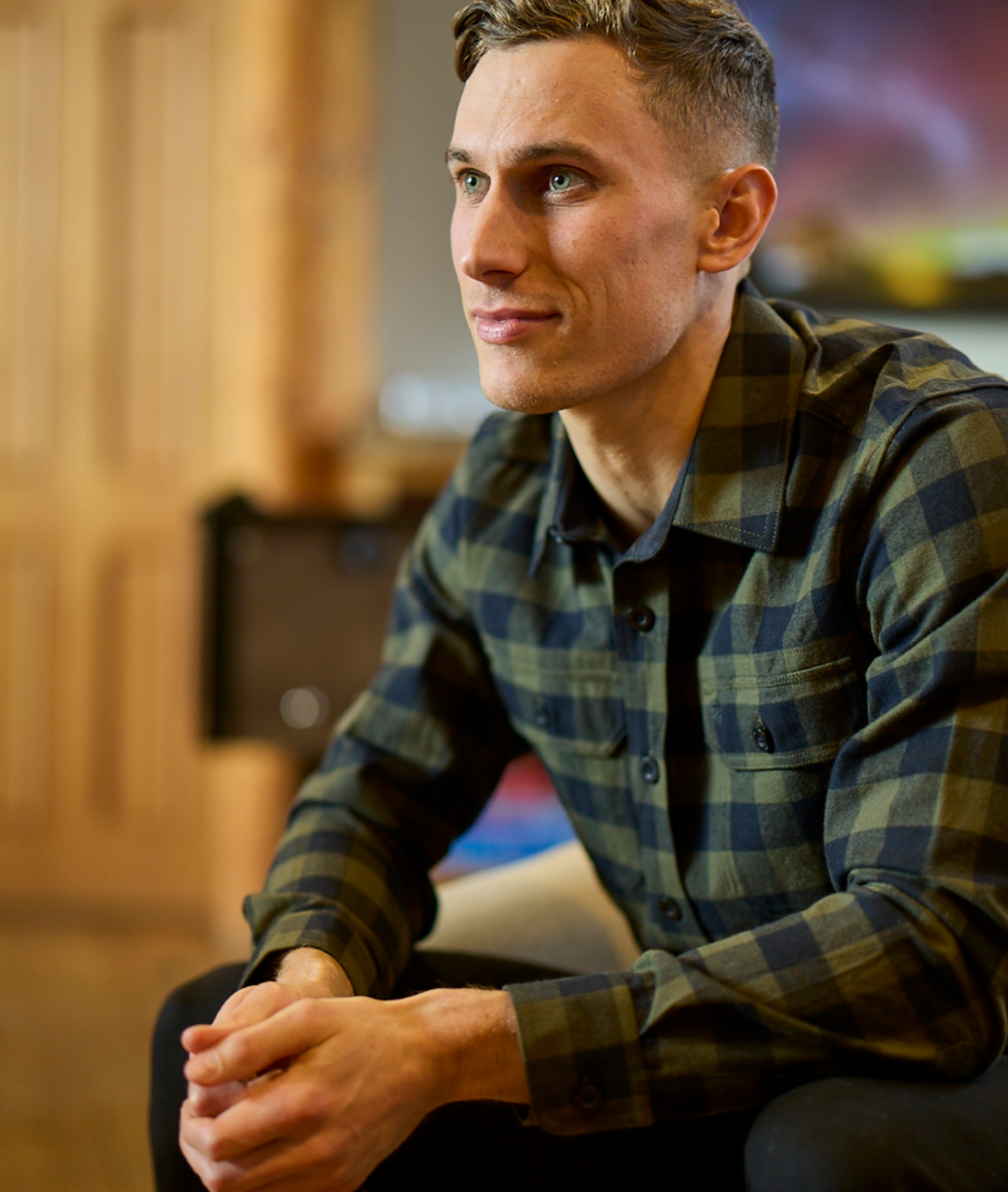 A man with short hair and blue eyes sits indoors, hands clasped and gazing thoughtfully aside, wearing the Swanndri Taranaki Tailor Shirt in green and black plaid. The softly blurred background enhances the scene.