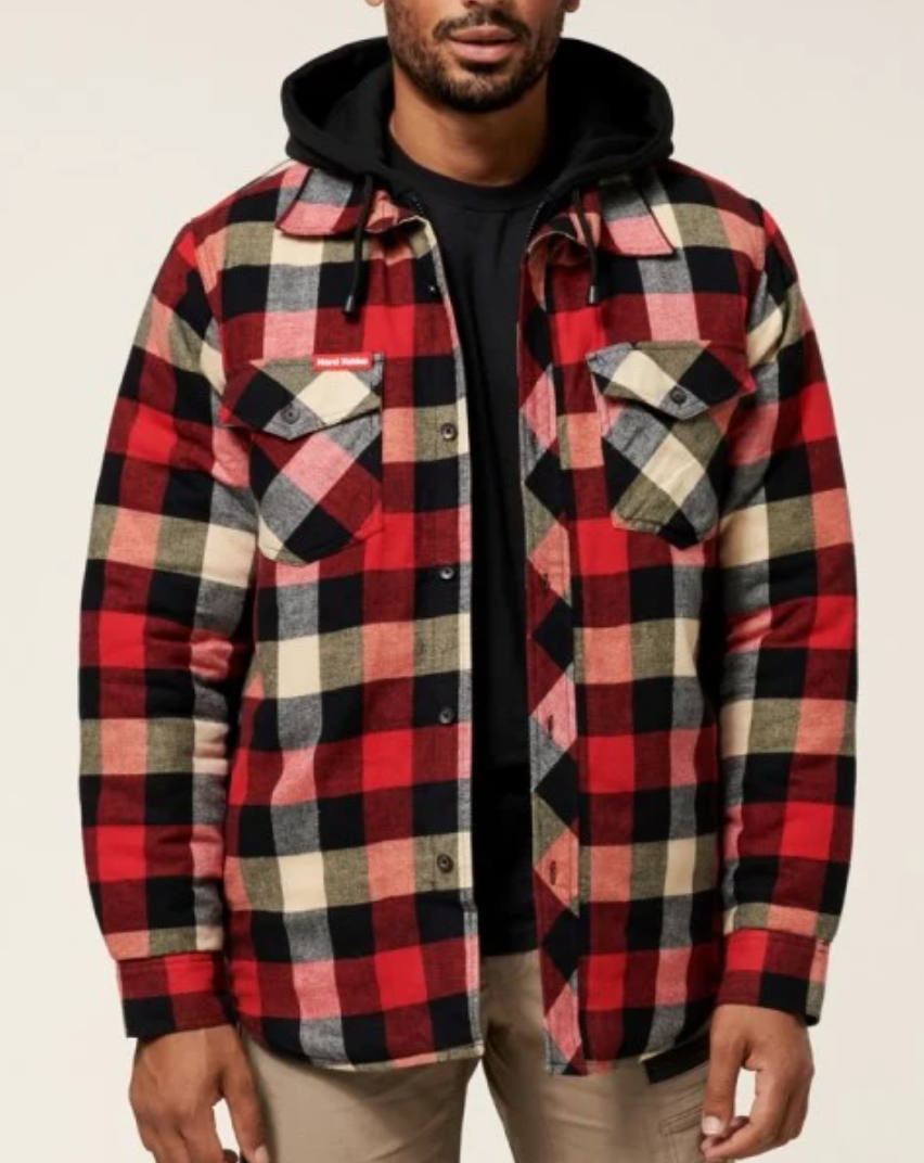 A man is wearing the Hard Yakka Quilted Flannelette Shacket, featuring a durable plaid pattern with two chest pockets and button closures, layered over a black hoodie. He has paired this ensemble with light brown pants. The scene is set against a plain, light-colored background.
