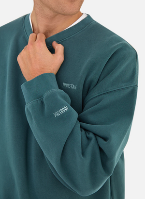 Wearing the "Industrie The Del Sur Sweat," featuring the logo on chest and sleeve, a person casually tugs at the neckline, adding a relaxed vibe to the oversized comfort jumper against a plain background.