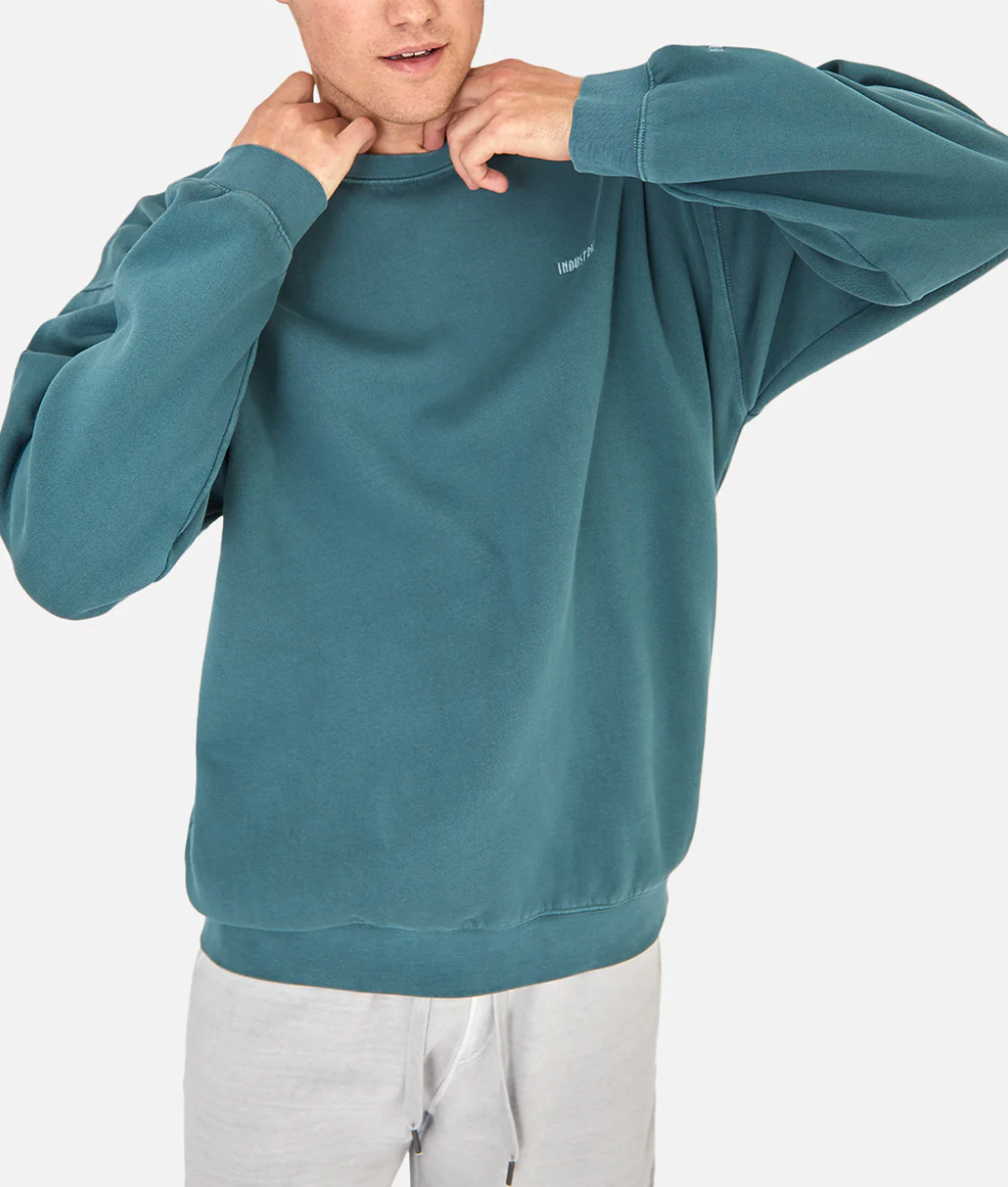 Someone wearing the Industrie The Del Sur Sweat in teal with an oversized fit and light gray pants adjusts the neckline of the jumper against a plain white background.