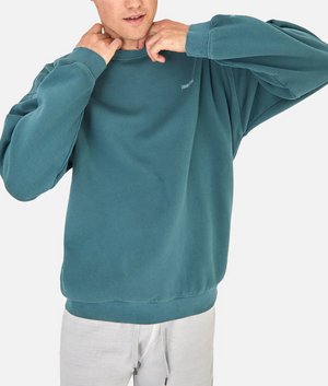 Someone wearing the Industrie The Del Sur Sweat in teal with an oversized fit and light gray pants adjusts the neckline of the jumper against a plain white background.
