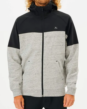 A person is wearing the Rip Curl Viral Anti Series Zip Thru Hood, which has a black top part and a light gray bottom part. This winter hoodie features a front zipper, side pockets, and small logos on the chest and left sleeve. The person, who has long curly hair, is standing against a plain white background.