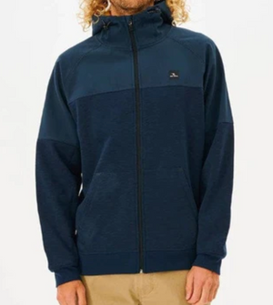 A person with curly hair is shown wearing the Rip Curl Viral Anti Series Zip Thru Hood in a dark blue color with a high collar. The Durable Water Resistant hoodie, featuring a small logo on the upper chest, is paired with beige pants against a plain white background.