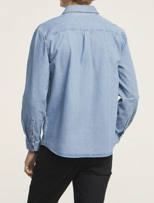 A person wearing a Wrangler Roadside Denim Shirt, in light blue and 100% cotton, paired with black pants, stands with their back to the camera. The shirt features long sleeves and buttoned cuffs against a plain white background.