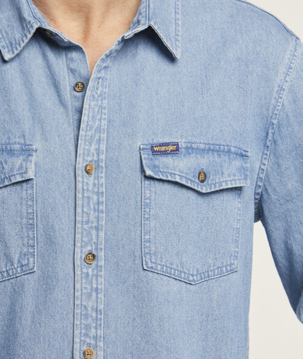 Someone wearing a light blue Wrangler Roadside Denim Shirt, made from 100% cotton. It has a collar, button-down front, two chest pockets with button flaps, and a small tag on the left pocket.