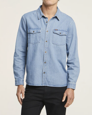 Someone in black pants dons a light blue Wrangler Roadside Denim Shirt, made of 100% cotton with long sleeves and two chest pockets. The plain off-white background emphasizes its classic style, though their head is not visible.