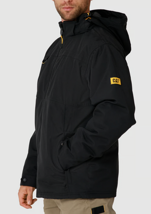 A man stands sideways with one hand in his pocket, dressed in khaki pants and the CAT Chinook Waterproof Jacket—black, featuring an adjustable hood and a yellow CAT logo patch on the sleeve.