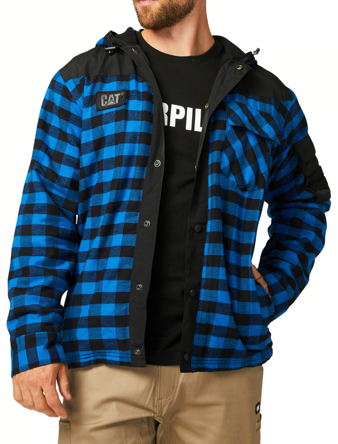A man wears the CAT Sequoia Shirt Jacket, a blue and black plaid jacket with durable overlays, black shoulder accents, a fleece-lined hood, and "CAT" logo, partially unbuttoned over a black Caterpillar T-shirt and tan pants.