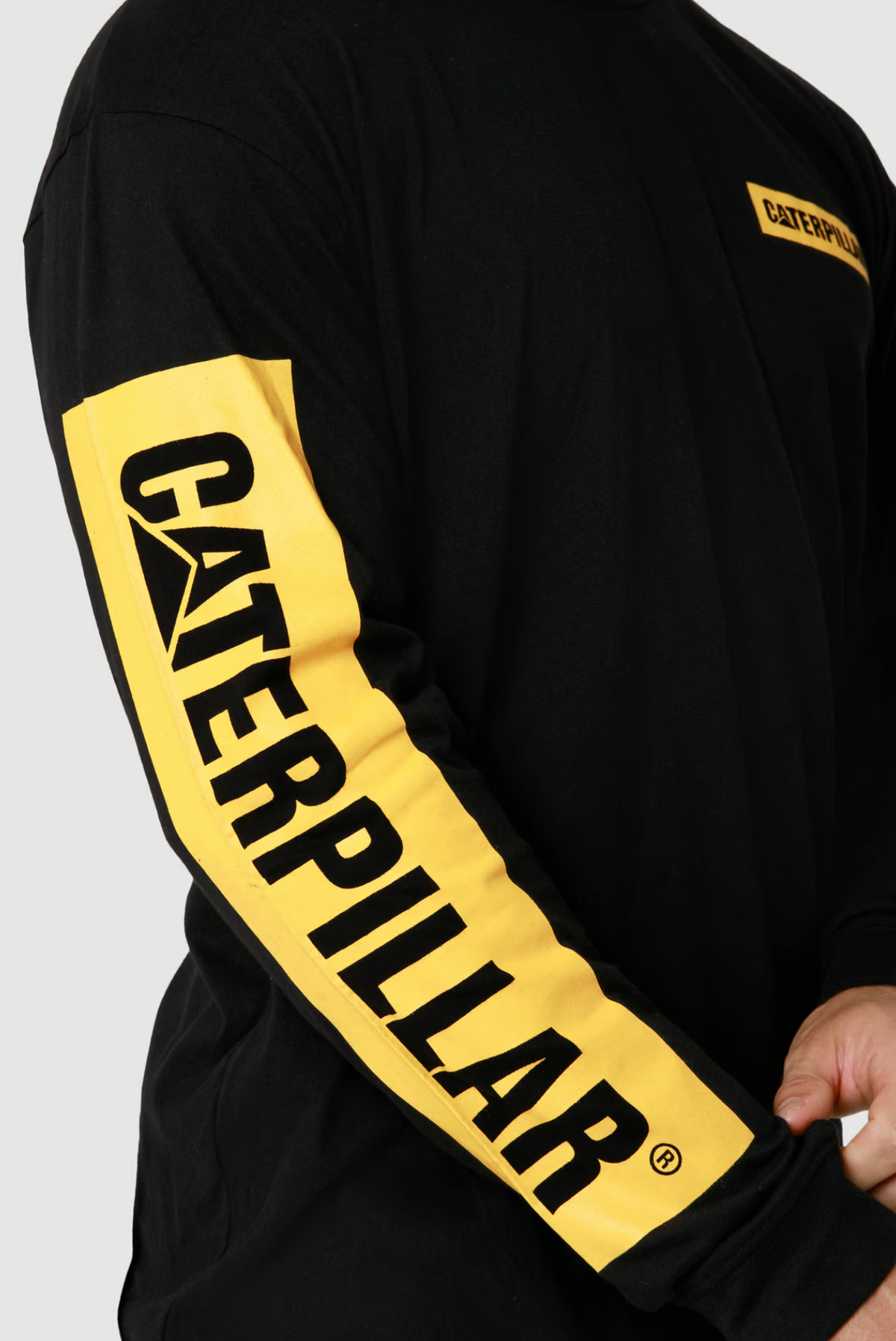 A person wears a CAT Icon Block Long Sleeve Tee, featuring a black crew neck, yellow sleeve panel with "CATERPILLAR" in bold black letters, and made from 100% cotton.