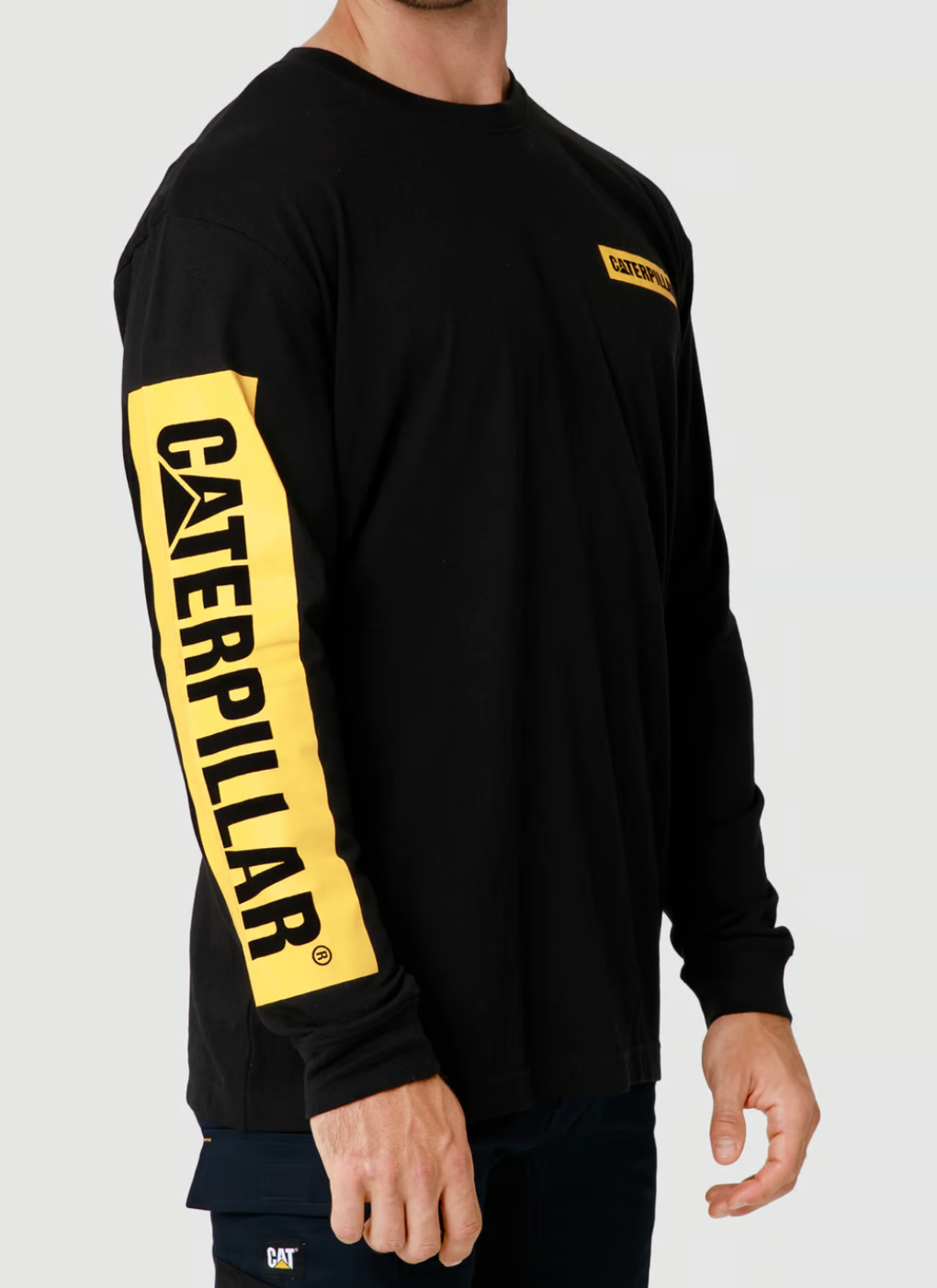A person wears the CAT Icon Block Long Sleeve Tee, a black shirt with "CATERPILLAR" in bold yellow on the left sleeve, 100% cotton with a crew neck, and a small yellow rectangle logo on the chest. The plain gray background accentuates its design.