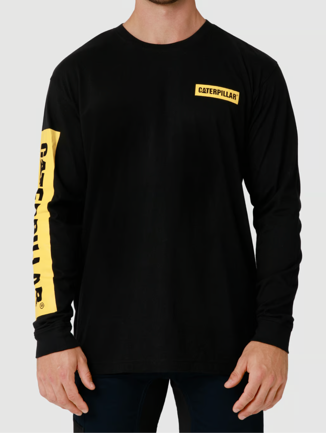 The man is wearing a CAT Icon Block Long Sleeve Tee in black, featuring a yellow logo patch on the chest and "Caterpillar" down the left sleeve. This crew neck shirt is made from 100% cotton for comfort and durability.