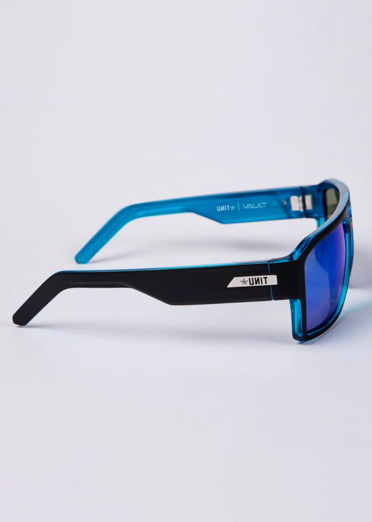 Side view of UNIT Vault Polarised Eyewear featuring sleek, blue-tinted lenses with UV protection and black arms. The modern translucent blue frame displays the brand name on the arm against a light gray backdrop.