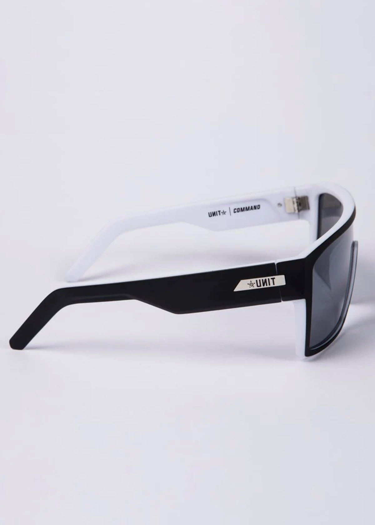 Side view of UNIT Command Polarised Eyewear: sporty black and white sunglasses with dark polarised lenses, thick lightweight frame, and gold UNIT logo on the arm, set against a plain light background.