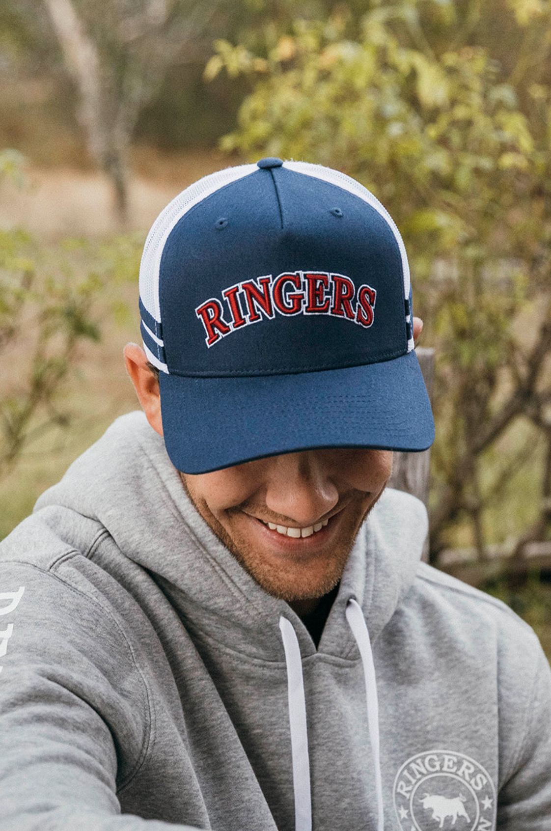 A smiling person in a gray hoodie and a Ringers Western Caleb Trucker Cap with "RINGERS" in red letters sits outdoors, greenery in the background.