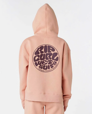 A person is wearing a light brown Rip Curl Wetsuit Icon Hood made from organic cotton, featuring a large circular "Rip Curl Wetsuits" logo on the back in brown text. The hoodie has cozy fleece lining, with the hood up as the person faces away from the camera.