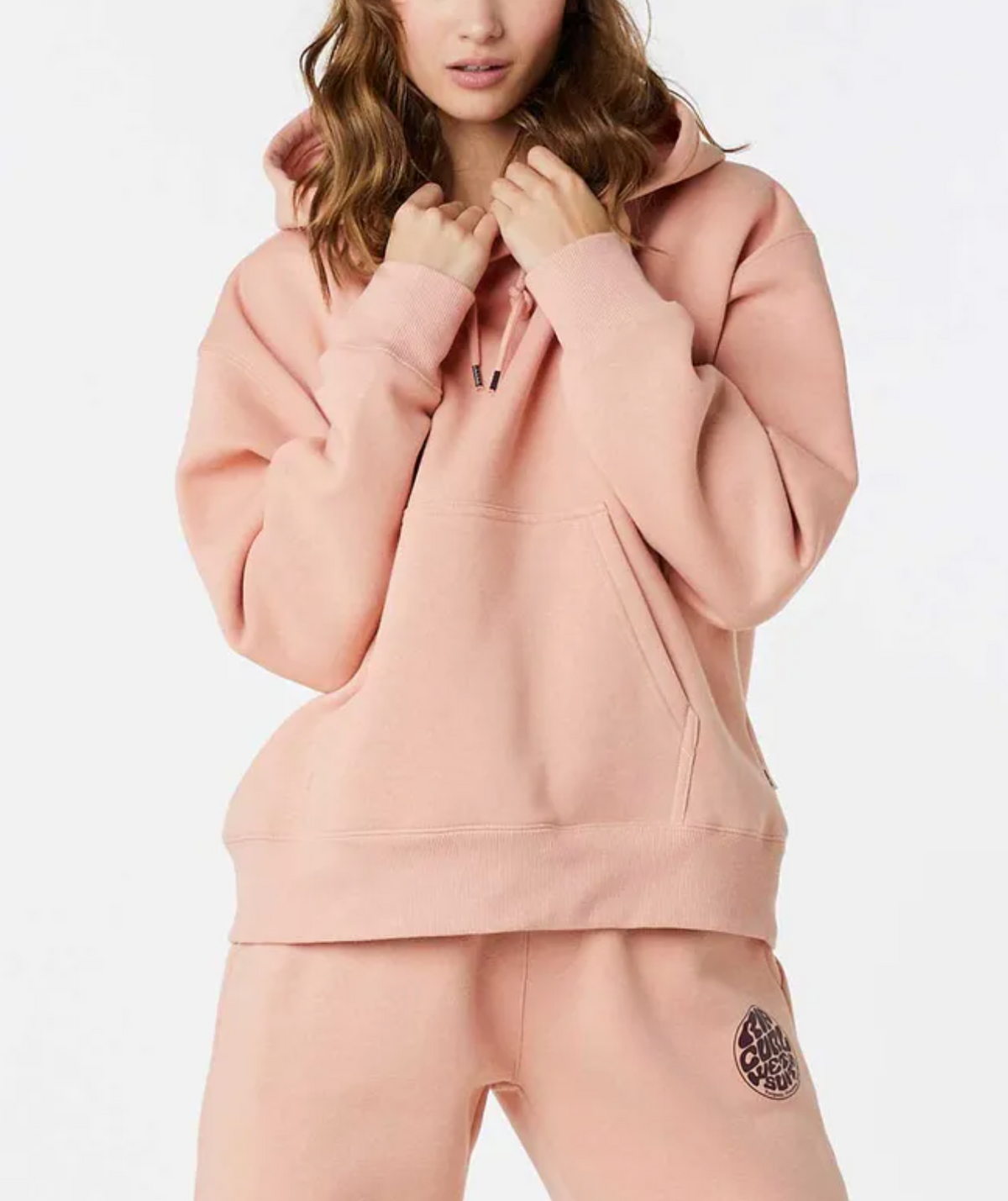 A person is wearing a pink Rip Curl Wetsuit Icon Hood made of organic cotton with matching joggers. The hoodie includes a drawstring, pouch pocket, and cozy fleece lining, while the joggers showcase a small circular logo on the left leg against a white background.