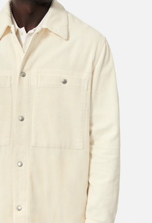 Wearing Industrie The Atmore Jacket, a cream corduroy jacket with stylish detailing and silver buttons, this person pairs it with a white shirt. Made of 100% cotton, it features two chest pockets and is photographed against a plain background.