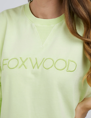 A close-up of a person in the Foxwood Simplified Crew, a light green cotton sweatshirt with Foxwood embroidery on the chest. The relaxed fit is paired with long, wavy brown hair draped over the right side.