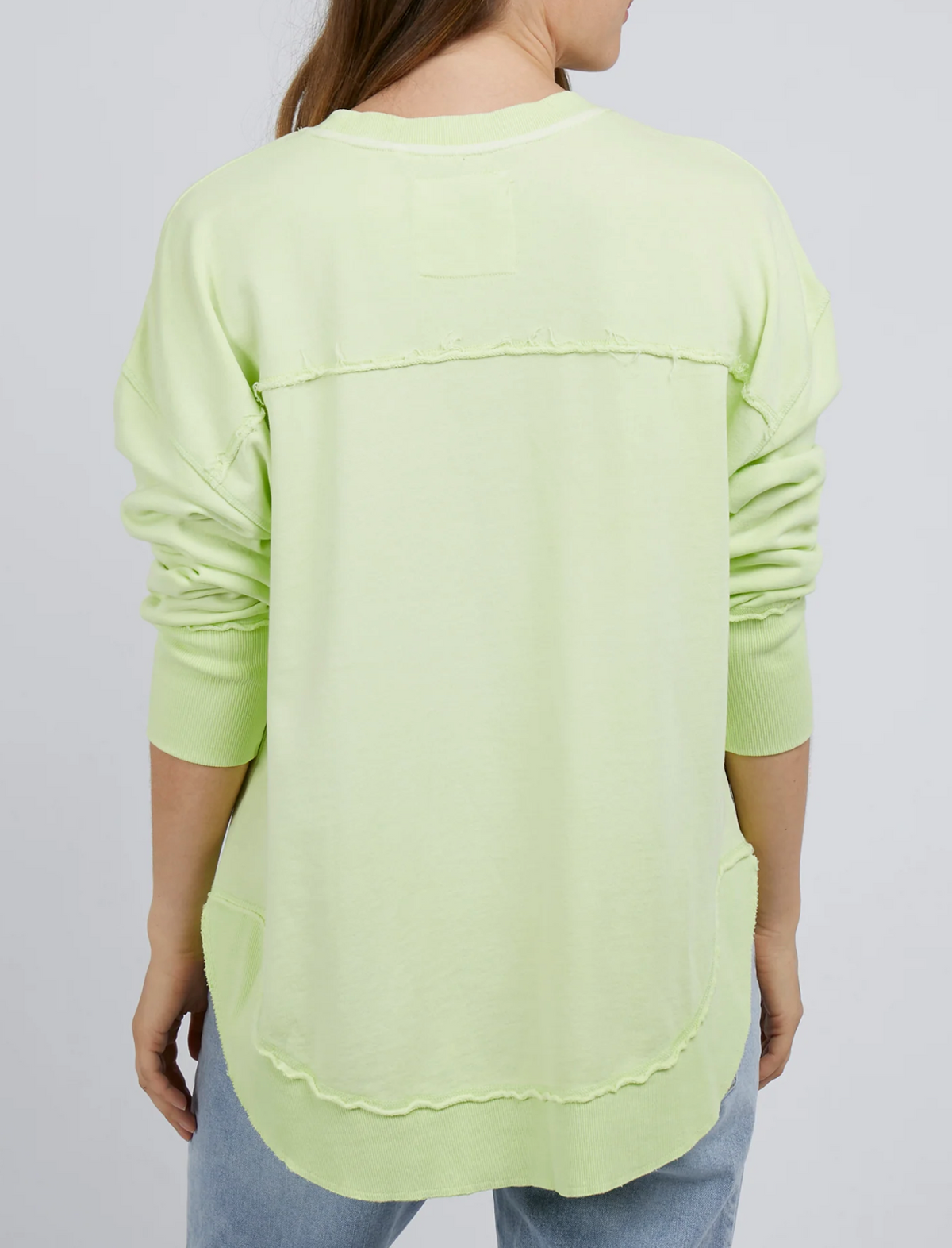 A person with long hair, seen from the back, is wearing the Foxwood Simplified Crew in light green—a relaxed fit long-sleeve cotton top with raw edge seams and ribbed cuffs—paired with light blue jeans.