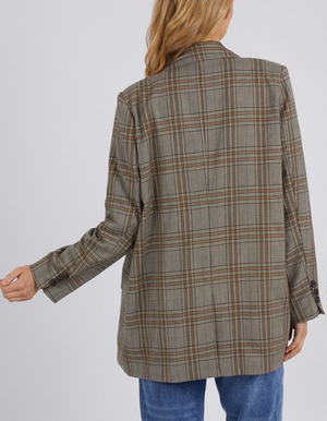 A person with long hair is seen from the back, wearing the oversized Foxwood Beatrix Blazer, a stylish brown and beige plaid design, paired with blue jeans. Their left hand is slightly extended.