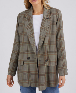 An individual sports the Foxwood Beatrix Blazer, an oversized plaid jacket in brown and gray tones, layered over a white shirt and blue jeans. This comfortable blazer boasts a double-breasted style with dark buttons and flap pockets, set against a plain gray backdrop.