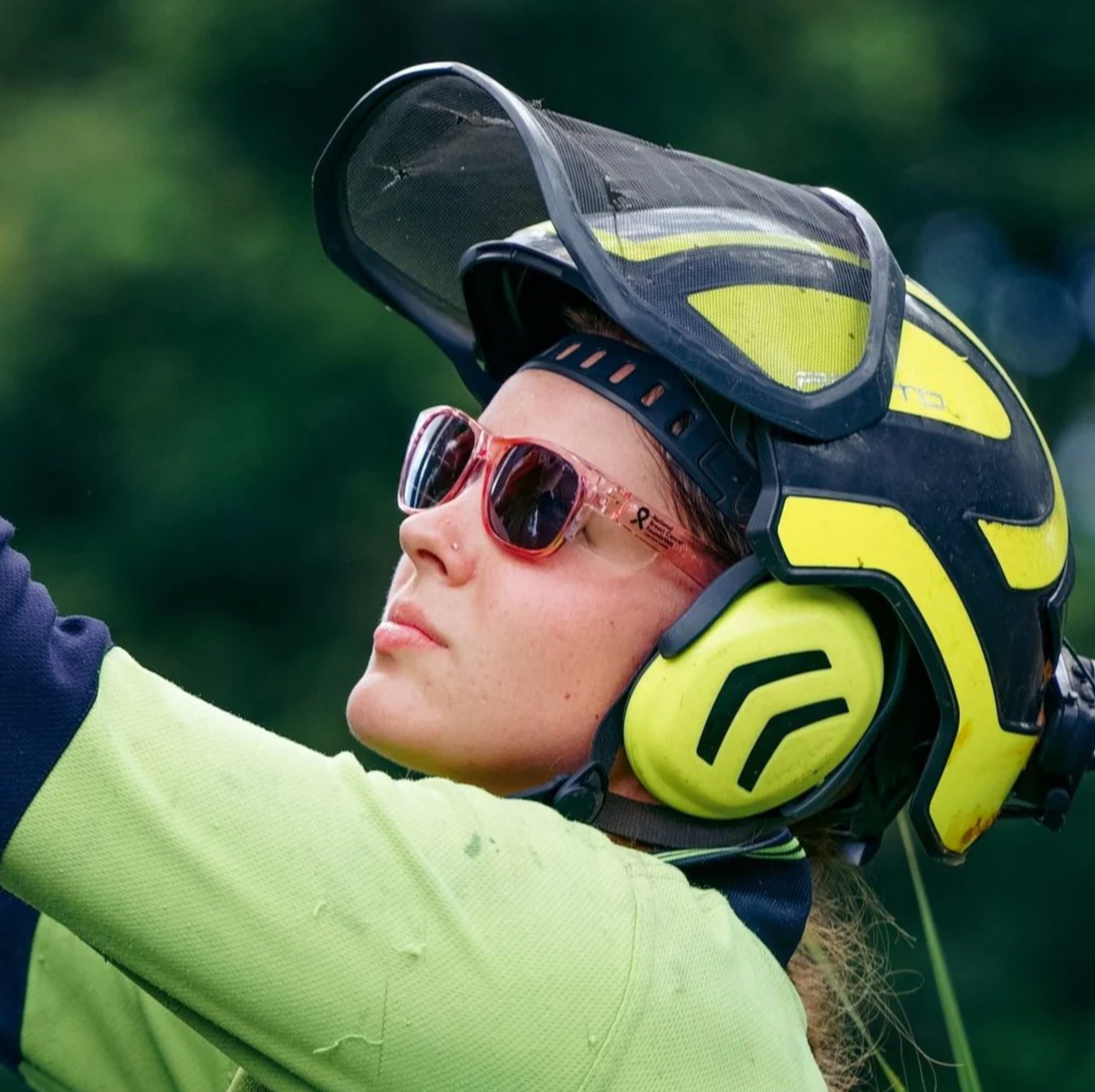Wearing safety gear, including a yellow and black helmet with a face shield, earmuffs, and SafeStyle Fusions Pink Frame Polarised Lens sunglasses, a person looks upward outdoors with blurred green foliage in the background.