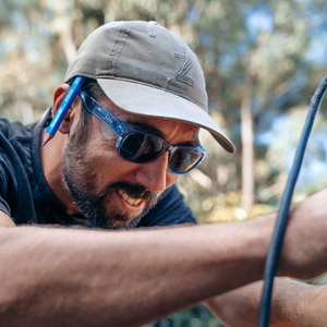 A man wearing SafeStyle Classics Blue Frame Tinted Lens sunglasses and a cap with a pen behind his ear is smiling while working outdoors, possibly on a DIY or gardening project, with trees and greenery blurred in the background.