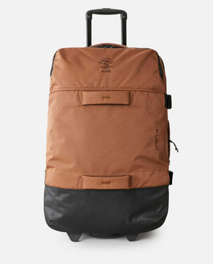 The Rip Curl F-Light Global 110L Searchers Travel Bag, in brown and black with a retractable handle, inline skate wheels, and a zippered front pocket, made from durable recycled CORDURA polyester, stands upright on a plain white background.