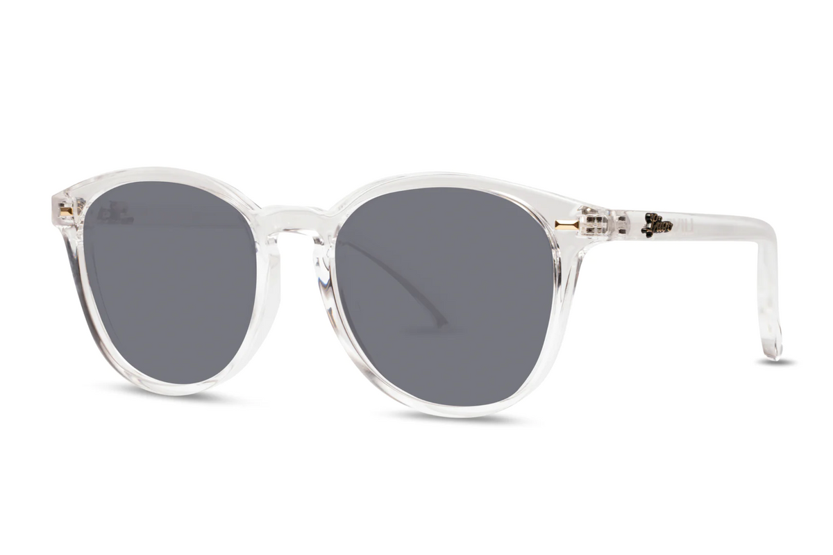 Liive Berawa Sunglasses feature clear round frames, dark gray UV-protective lenses, and a minimal modern design. Crafted from lightweight polycarbonate, they are presented on a plain white background.