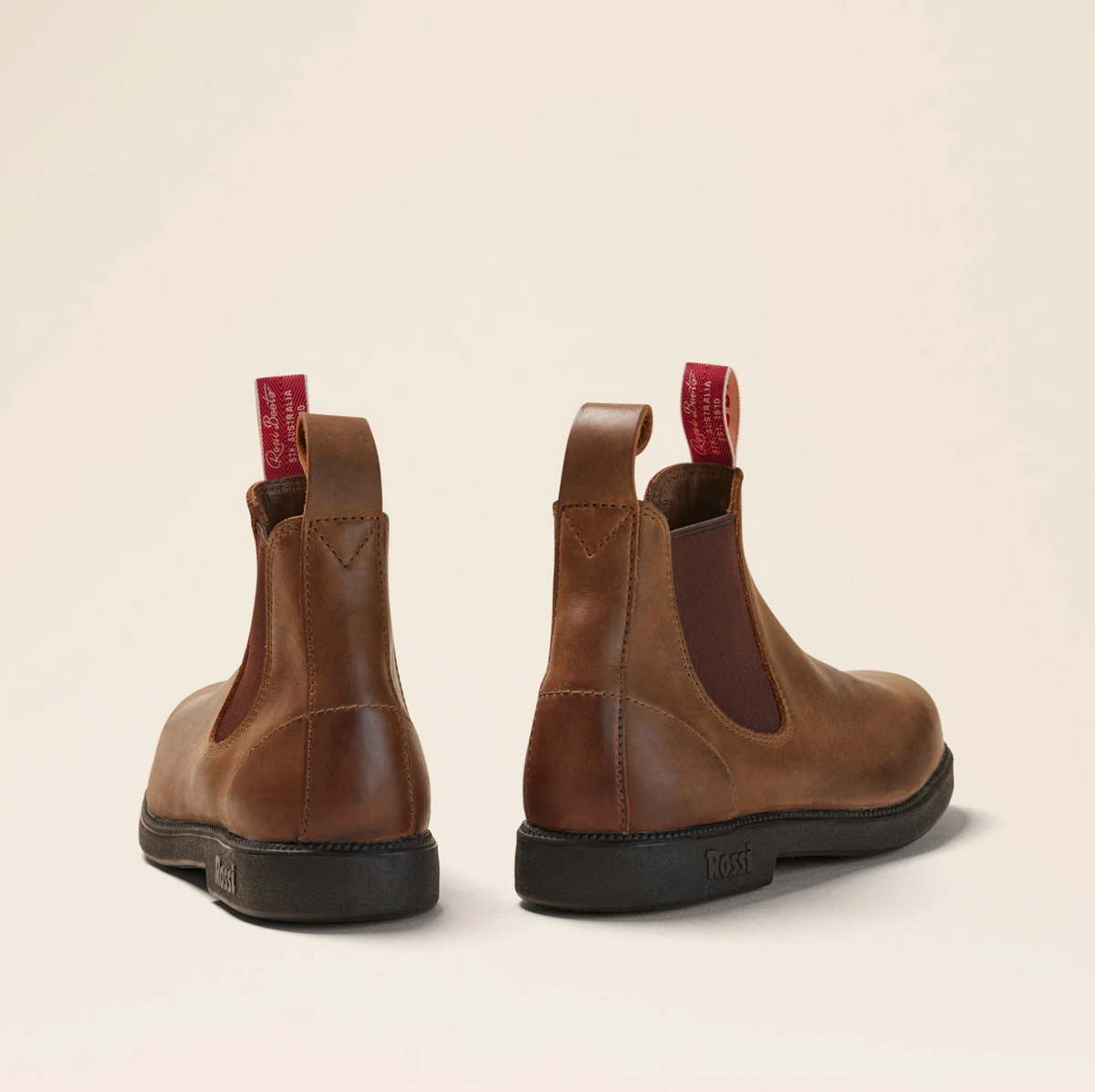 The Rossi Booma 607 Elastic Sided Boot features a pair of brown Chelsea boots made from Full Grain Australian Leather and equipped with black, slip-resistant soles. They are designed with red pull tabs on the back and elastic side panels for easy wear. Positioned on a light beige background, the boots are displayed showcasing both the side and back views.