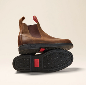 The Rossi Booma 607 Elastic Sided Boot is a pair of brown Full Grain Australian Leather Chelsea boots, featuring pull tabs. One boot stands upright while the other lies on its side, highlighting the slip-resistant black rubber sole with a red tag and circular tread pattern. The background is light beige.