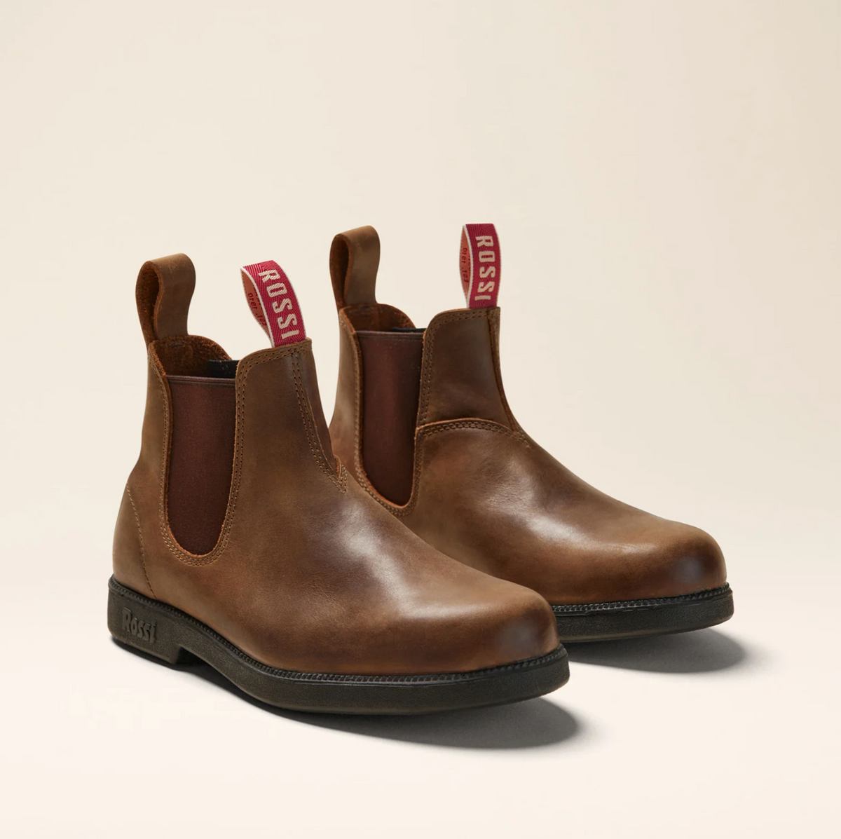 The Rossi Booma 607 Elastic Sided Boot, made from Full Grain Australian Leather in brown, is showcased on a neutral background. These Chelsea boots feature elastic side panels and pull tabs labeled "ROSSI," as well as slip-resistant black soles. The boots boast a smooth finish and a sleek, classic design ideal for both work and casual wear.